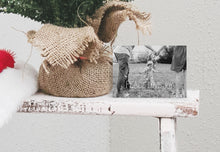 Load image into Gallery viewer, A photo of a one-sided Christmas card showing the front of the card standing up next to the bottom of a small Christmas tree with the base wrapped in burlap. The card features a photo with the words “Peace Love Joy” over the photo and a space to put your family name below. There are illustrated modern trees on the bottom. 