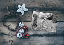 Load image into Gallery viewer, A photo of a one-sided Christmas card showing the front and back of the card laying on a wood surface. To the left of the cards are some ornaments. The card features a photo with the words “Peace Love Joy” over the photo and a space to put your family name below. There are illustrated modern trees on the bottom. 