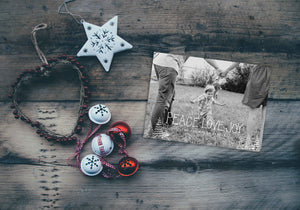 A photo of a one-sided Christmas card showing the front and back of the card laying on a wood surface. To the left of the cards are some ornaments. The card features a photo with the words “Peace Love Joy” over the photo and a space to put your family name below. There are illustrated modern trees on the bottom. 