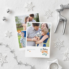 Load image into Gallery viewer, A photo of a double-sided Christmas card showing the front and back of the card laying on a white surface. Around the two sides of the card are surrounded with Christmas items. The front of the card features a photo with the words “Peace Love joy” to the bottom right and space to the bottom left to put your family names. The words feature some modern illustrated flowers around it. The back of the card features three photos with modern illustrated flowers.