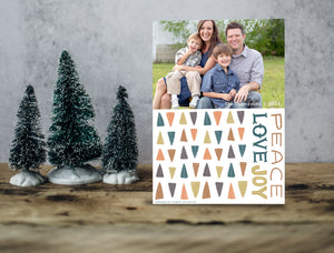 A photo of a one-sided Christmas card showing the front of the card standing up with three small Christmas trees next to it. The photo card features one photo with illustrated modern Christmas trees below. The words “Peace Love Joy” are to the left of the tree pattern. 