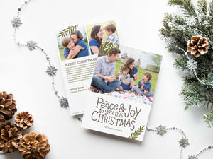 A photo of a double-sided Christmas card showing the front and back of the card laying on a white surface. Around the two sides of the card are pine cones, pine needles and a string of silver snowflake garland. The front of the card features a photo on the top portion and the words “Peace & Joy to You This Christmas” with space to put your family name below. The back side of the card features two photos with space to write a yearly update. 