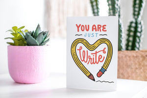 A greeting card featured standing up on a white tabletop with a pink plant pot in the background and some succulents in the pot. There’s a woven basket in the background with a cactus inside. The card features the words “You are just write”with an illustrated pencil in the shape of a heart.