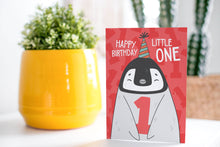 Load image into Gallery viewer, A greeting card standing up on a table with a plant in a yellow pot behind it. The card features an illustrated penguin with a party hat on and holding the number one and reads &quot;Happy birthday little one.&quot;