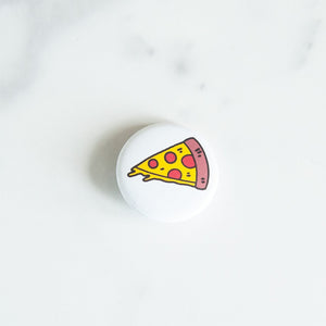 A button pin on a white tabletop. The pin features an illustration of a slice of pizza.