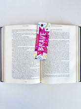 Load image into Gallery viewer, A bookmark with the word &quot;brave&quot; in white within a star-like pink shape with fun, sketchy elemts around. The bookmark is placed inside a book. 