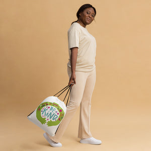 A woman walking while holding a white drawstring bag. The drawstring bag has the words "Be Kind" with green illustrated hands hugging the design with sketchy illustrations. 