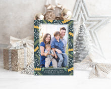 Load image into Gallery viewer, A photo of a one-sided Christmas card showing the front of the card standing up with Christmas items behind the card. There’s a sparkly star, glitter wrapped gift, ribbon and ornaments with a small silver tree around the photo card. The photo card features one photo with a border of illustrated gold crowns, and green and white illustrated leaves. The bottom of the card reads “Celebrate the Prince of Peace, The Thompsons.”