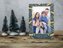 Load image into Gallery viewer, A photo of a one-sided Christmas card showing the front of the card standing up with three small Christmas trees next to it. The photo card features one photo with a border of illustrated gold crowns, and green and white illustrated leaves. The bottom of the card reads “Celebrate the Prince of Peace, The Thompsons.”