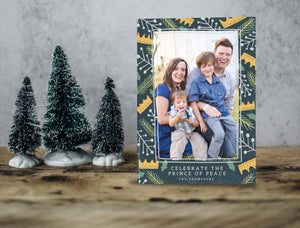 A photo of a one-sided Christmas card showing the front of the card standing up with three small Christmas trees next to it. The photo card features one photo with a border of illustrated gold crowns, and green and white illustrated leaves. The bottom of the card reads “Celebrate the Prince of Peace, The Thompsons.”