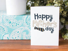 Load image into Gallery viewer, A greeting card is on a table top with a present in blue wrapping paper in the background. On top of the present is a candle and some greenery from a plant too. The card features the words  “Happy Father’s  Day” with a striped tie on the bottom of the words. 