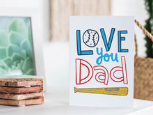 Load image into Gallery viewer, A greeting card featured standing up on a white tabletop with a framed photo of a succulent in the background and a stack of wooden coasters. There’s a woven basket in the background with a cactus inside. The card features the words “Love You Dad” with an illustrated baseball as the “O” of love and a baseball bat featured at the bottom of the words. 