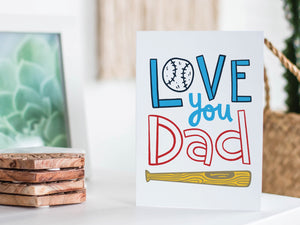 A greeting card featured standing up on a white tabletop with a framed photo of a succulent in the background and a stack of wooden coasters. There’s a woven basket in the background with a cactus inside. The card features the words “Love You Dad” with an illustrated baseball as the “O” of love and a baseball bat featured at the bottom of the words. 
