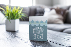 A greeting card featured on a black, wood coffee table. There’s a white planter in the background with a green plant. There’s also a gray sofa in the background with a white pillow. The card features the words “When you go through deep waters, I will be with you.”