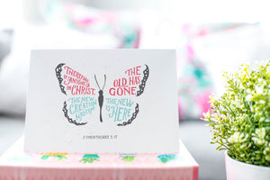 A greeting card is on a table top with a gift in pink wrapping paper. Next to the gift is a white plant pot with a green plant. The card features the words “Therefore if anyone is in Christ, the new creation has come. The old has gone. The new is here. 2 Corinthians 5:17.” The card design features an illustrated butterfly. 