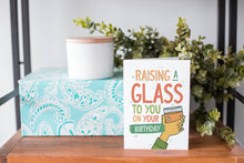 Load image into Gallery viewer, A greeting card is on a table top with a present in blue wrapping paper in the background. On top of the present is a candle and some greenery from a plant too. The card features the words “Raising a glass to you on your birthday” with an illustrated hand raising a glass.