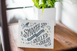 A photo of a card featured on a tabletop next to a white planter filled with a green plant. ​​The card features the words “A smooth sea never made a skilled sailor.”