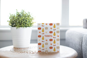 A greeting card is featured on round coffee table with a green plant and sofa in the background. The card features the word "Thankful" with a pattern of illustrated pumpkins and leaves behind the word. 