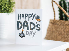 Load image into Gallery viewer, A greeting card is featured on a white tabletop with a white planter in the background with a green plant. There’s a woven basket in the background with a cactus inside. The card features the words “Happy Dad’s Day” with an illustrated game controller and hat. 