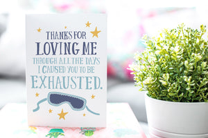A photo of a card featured on a tabletop next to a white planter filled with a green plant. ​​The card features the words “Thanks for Loving Me All the Days I Caused You to be Exhausted.”
