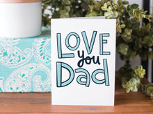 Load image into Gallery viewer, A greeting card is on a table top with a present in blue wrapping paper in the background. On top of the present is a candle and some greenery from a plant too. The card features the words  “Love You Dad” in simple typography. 