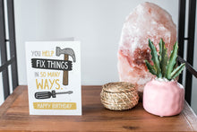 Load image into Gallery viewer, A card on a wood tabletop and on the right side of the card is a woven basket, a pink plant pot with a cactus in it and a pink crystal rock. The card features the words “You help fix things in so many ways. Happy Birthday” featuring an illustrated hammer.
