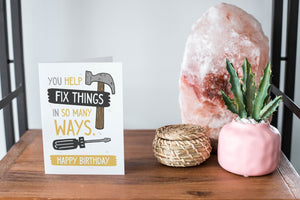 A card on a wood tabletop and on the right side of the card is a woven basket, a pink plant pot with a cactus in it and a pink crystal rock. The card features the words “You help fix things in so many ways. Happy Birthday” featuring an illustrated hammer.
