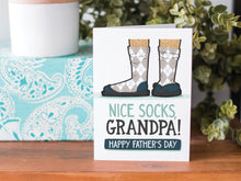 Load image into Gallery viewer, A greeting card is on a table top with a present in blue wrapping paper in the background. On top of the present is a candle and some greenery from a plant too. The card features the words  “Nice Socks Grandpa, Happy Father’s Day” with an illustrated of legs with patterned socks and shoes. 
