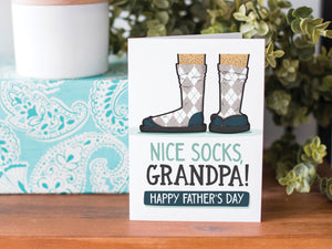 A greeting card is on a table top with a present in blue wrapping paper in the background. On top of the present is a candle and some greenery from a plant too. The card features the words  “Nice Socks Grandpa, Happy Father’s Day” with an illustrated of legs with patterned socks and shoes. 