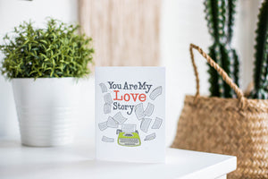A greeting card is featured on a white tabletop with a white planter in the background with a green plant. There’s a woven basket in the background with a cactus inside. The card features the words “You are my love story” with an illustrated typewriter and scattered papers.