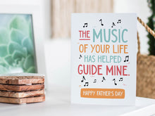 Load image into Gallery viewer, A greeting card featured standing up on a white tabletop with a framed photo of a succulent in the background and a stack of wooden coasters. There’s a woven basket in the background with a cactus inside. The card features the words “The music of your life has helped guide mine. Happy Father&#39;s Day.” 