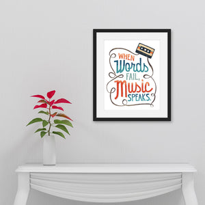 Artwork is featured in a black frame on a white wall. The art is hanging above a white hallway table with a black on top of it. The artwork features hand drawn lettering with the phrase "When words fail, music speaks." In the upper corner of the words an illustrated cassette tape is featured.