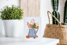 Load image into Gallery viewer, A greeting card featured standing up on a white tabletop with a white plant pot with a green plant. There’s a woven basket in the background with a cactus inside. The card features the words &quot;May Your Table be Filled with Loved Ones&quot; with the words inside an illustrated watering can with leaves coming out of the top.