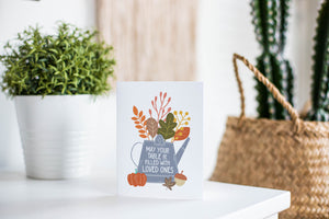 A greeting card featured standing up on a white tabletop with a white plant pot with a green plant. There’s a woven basket in the background with a cactus inside. The card features the words "May Your Table be Filled with Loved Ones" with the words inside an illustrated watering can with leaves coming out of the top.