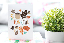 Load image into Gallery viewer, A greeting card is featured on pink wrapped gift with a green plant in the background. The card features illustrated lettering reading “Thankful&quot; with illustrated leaves and an acorn around the word.