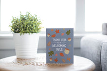 Load image into Gallery viewer, A greeting card is featured on round coffee table with a green plant and sofa in the background. The card features the words &quot;Thank You for a Welcoming Table&quot; with illustrated leaves and acorns around the words. 