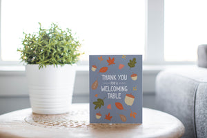 A greeting card is featured on round coffee table with a green plant and sofa in the background. The card features the words "Thank You for a Welcoming Table" with illustrated leaves and acorns around the words. 