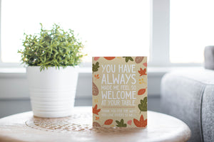 A greeting card is featured on round coffee table with a green plant and sofa in the background. The card features the words "You have always made me feel so welcome at your table. Thank you for the ways You love others so well" with illustrated leaves surrounding the words.