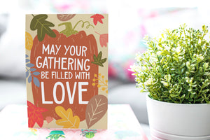 A greeting card is featured on pink wrapped gift with a green plant in the background. The card features illustrated lettering reading "May Your Gathering Be Filled with Love" with the words inside an illustrated pumpkin with leaves surrounding the pumpkin. 