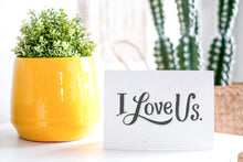Load image into Gallery viewer, A greeting card is on a table top with a yellow plant pot and a green plant inside. The card features the words “I Love Us.”