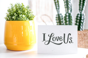 A greeting card is on a table top with a yellow plant pot and a green plant inside. The card features the words “I Love Us.”