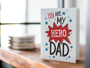 A card on a wood tabletop with an object in the background that is out of focus. The card features the words "You are my hero Dad.” 