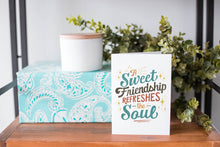 Load image into Gallery viewer, A greeting card is on a table top with a present in blue wrapping paper in the background. On top of the present is a candle and some greenery from a plant too. The card features the words “A sweet friendship refreshes the soul.”