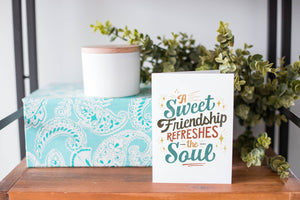 A greeting card is on a table top with a present in blue wrapping paper in the background. On top of the present is a candle and some greenery from a plant too. The card features the words “A sweet friendship refreshes the soul.”