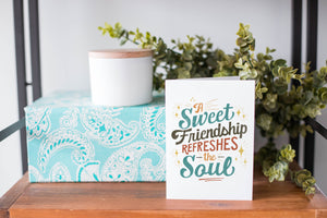 A white greeting card sitting on a a table top with a gift and greenery behind it. The card has the words "A Sweet Friendship Refreshes the Soul."