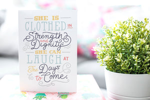 A greeting card is on a table top with a gift in pink wrapping paper. Next to the gift is a white plant pot with a green plant. The card features the words "She is clothed in strength and dignity; she can laugh at the days to come." 
