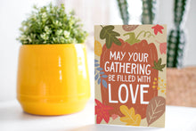 Load image into Gallery viewer, A greeting card is on a table top with a yellow plant pot and a green plant inside. The card features the words &quot;May Your Gathering Be Filled with Love&quot; with the words inside an illustrated pumpkin with leaves surrounding the pumpkin. 