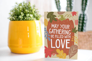 A greeting card is on a table top with a yellow plant pot and a green plant inside. The card features the words "May Your Gathering Be Filled with Love" with the words inside an illustrated pumpkin with leaves surrounding the pumpkin. 