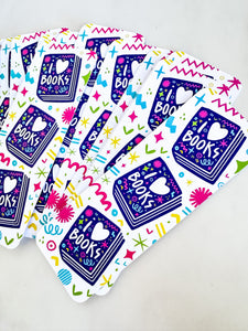 BookmarkS on a tabletop. The bookmarks feature the words "i love books" with love shown shaped as a heart. The words are placed inside an illustrated book. 