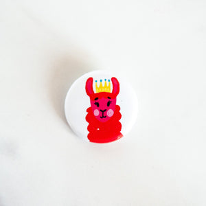 A buttons on a white tabletop. The button features a red illustrated llama with crown on. 